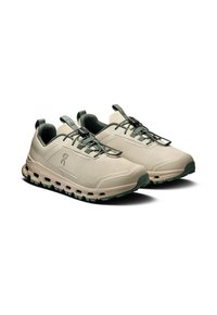 Beige Sport-Schuhe mit grau-grünen Akzenten, Obermaterial aus strukturiertem Stoff und flexibler Sohle mit Luftpolstern zur Dämpfung. Schnürsenkel mit Schnappverschluss.