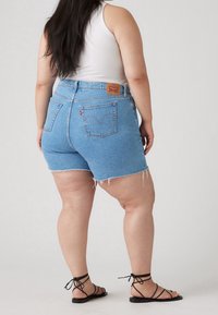 Shorts en denim bleu clair avec bords effilochés, cinq poches et un patch logo en cuir à l'arrière. Portés avec des sandales noires à lanières.