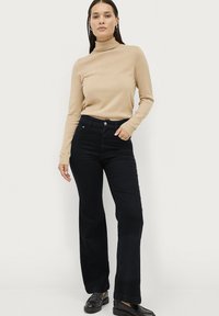 Beige Rollkragenpullover und schwarze weit geschnittene Hose, getragen mit schwarzen Slippern. Der Pullover hat Rippstrick; die Hose hat einen hohen Bund und Taschen.