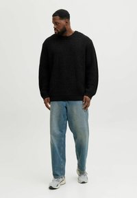 Mann mit kurzen lockigen Haaren und Bart, der einen schwarzen Strickpullover, lockere blaue Jeans und graue Turnschuhe trägt und nach links schaut, vor einem weißen Hintergrund.