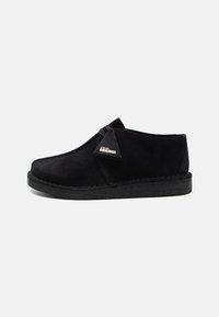 Black suede shoe with a rounded toe, a minimalistic design, and a crepe sole. Features a small brand tag on the side.