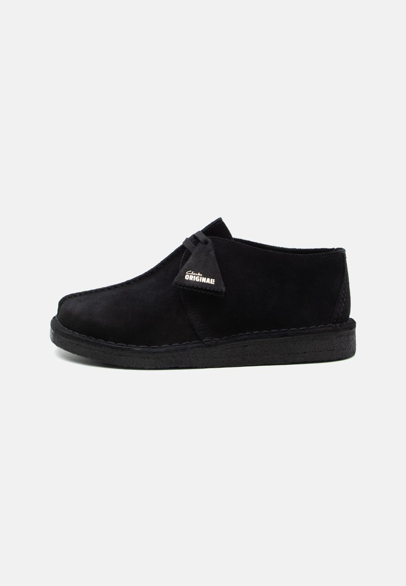 Black suede shoe with a rounded toe, a minimalistic design, and a crepe sole. Features a small brand tag on the side.