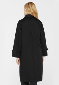 Eine blonde Person, die einen langen schwarzen Mantel trägt, von hinten vor einem schlichten weißen Hintergrund gesehen.