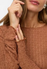 Brązowa, teksturowana bluzka z długim rękawem i postrzępionymi mankietami. Tkanina ma delikatny wzór, podkreślający miękki, lekki wygląd.