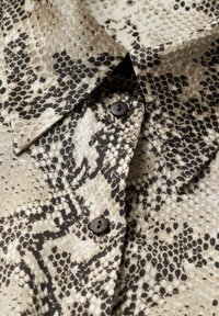 Yllä oleva kauluspaita on valmistettu beigestä ja mustasta käärmeennahkakuosisesta kankaasta, ja sen kauluksessa on kaksi mustaa nappia kiinnitettynä.