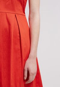 Braccio di donna appoggiato accanto a un vestito rosso a pieghe su uno sfondo chiaro e semplice.