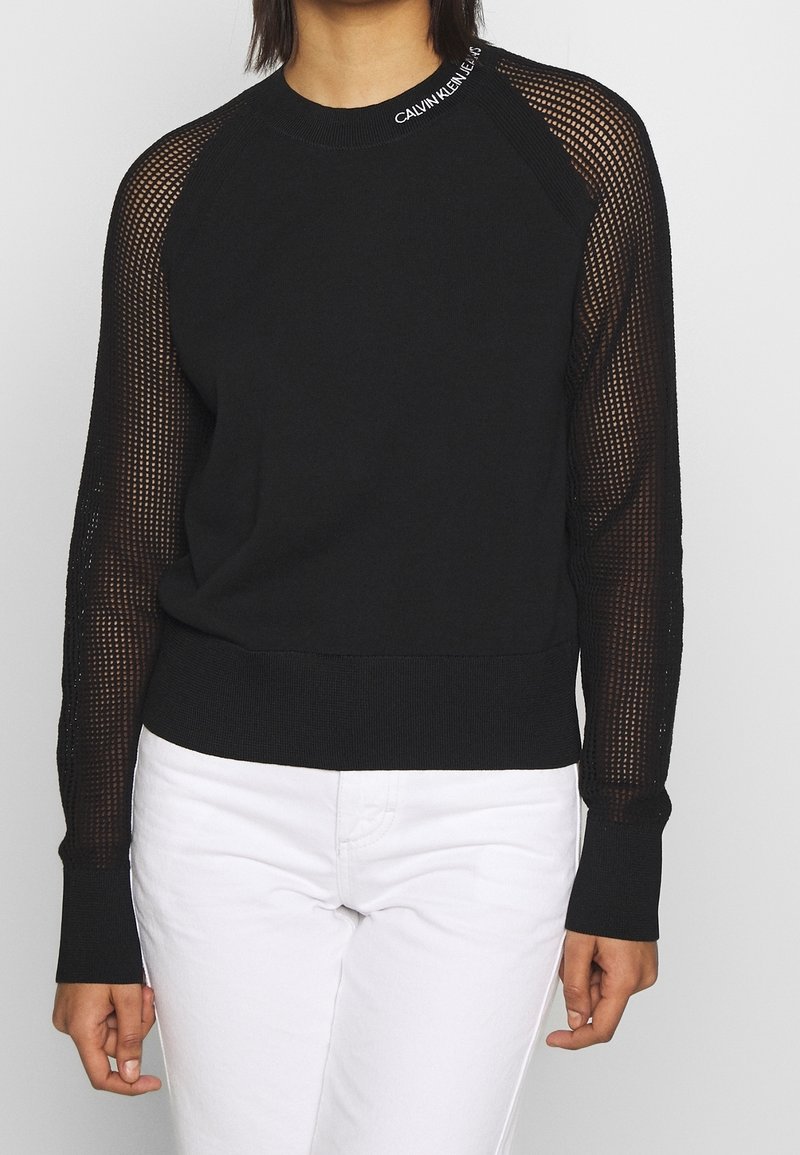 Czarny sweter z siateczkowymi rękawami, ściągaczami przy mankietach i dekolcie z logo "CALVIN KLEIN JEANS". Gładki główny materiał oraz teksturowane akcenty.