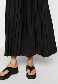 Gonna maxi plissettata nera abbinata a sandali flatform neri con un design a strappo a forma di V. I sandali hanno una suola spessa per supporto.