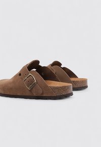 Zuecos de gamuza marrón con una hebilla metálica ajustable, plantilla de corcho y suela de goma texturizada, con un diseño de espalda abierta.