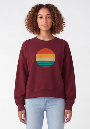 Jeune femme aux cheveux bouclés portant un sweat-shirt bordeaux avec un graphisme cercle coucher de soleil rétro et un jean bleu clair, debout sur fond blanc.