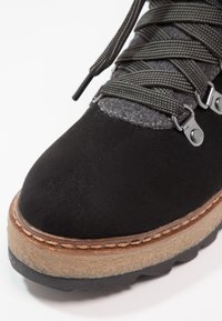 Botín exterior de ante negro con puño de tela gris, herrajes metálicos y suela marrón claro. Presenta una parte inferior texturizada y cordones trenzados.