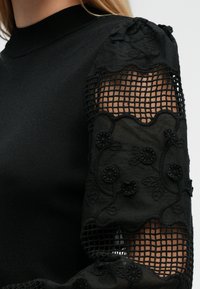 Top nero con corpo aderente e maniche in pizzo ricamato voluminose, con motivi floreali e design a rete a griglia.