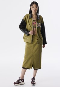 Veste utilitaire vert olive avec des manches en cuir noir, crop top avec des lettres rouges, jupe olive jusqu’aux genoux et baskets bourgogne.