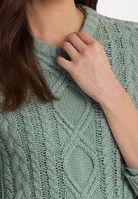 Ljusgrön stickad tröja med en texturerad kabeldesign och en bred halsringning. Tyget verkar tjockt och mysigt.