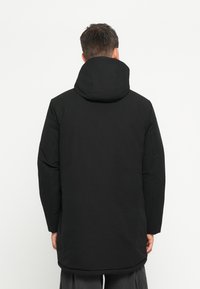 Zwarte lange jas met een capuchon, zichtbare stiksels en een gladde textuur. De mouwen zijn iets pufachtig met een recht gesneden silhouet.