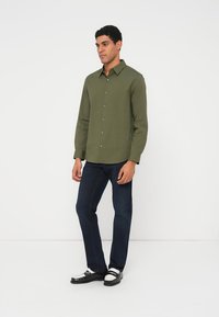 Olivzaļš krekls ar garām piedurknēm un pogu aizdari, kombinēts ar tumši zilām džinsām un melni baltām kurpēm. Gluda tekstūra, ikdienas piegriezums.