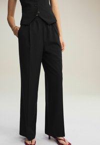 Schwarze maßgeschneiderte Hose mit weitem Beinschnitt, ausgestattet mit einem elastischen Bund und einer glatten Textur. Kombiniert mit roten, hohen Sandalen.