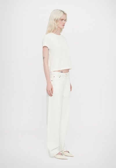 T-shirt blanc court à manches courtes, associé à un pantalon blanc taille haute ample. Les chaussures sont des ballerines de couleur claire.