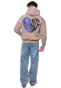 Beige Kapuzenpulli mit einem Herz-Graphic in Blau und Schwarz mit gewellten Mustern. Kombiniert mit hellblauen, weiten Jeans.