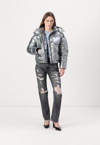 Sølv metallic pufferjakke med hette, glidelås foran, og lommepatch, parret med slitte svarte jeans og svarte høye hæler.