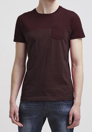 Voksen iført en kortærmet T-shirt i burgunder og mørkebrun farveblok med brystlomme og blå jeans.