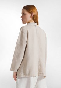 Giacca in lino beige chiaro con cucitura posteriore e dettaglio di cucitura sottile. Presenta un collo con revers e una silhouette morbida e fluente.