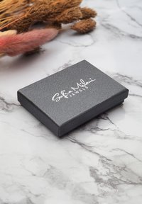 Grå rektangulär smyckesask med texturerad yta och silverlogotyp, placerad på en marmorbakgrund med torkade blommor i närheten.