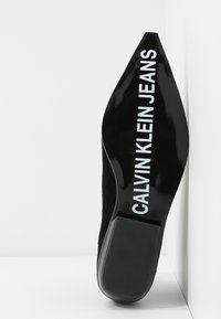 Zapato de tacón alto negro con punta afilada, material de ante y un tacón negro brillante que presenta "CALVIN KLEIN JEANS" impreso en blanco.