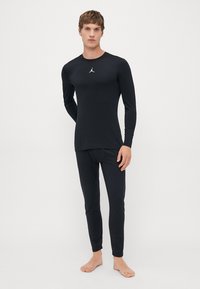 Man in een zwart langemouwen shirt en strakzittende broek met een klein wit logo op de borst, staande op blote voeten tegen een effen achtergrond.