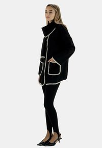 Cappotto nero con bordi bianchi, colletto alto, due tasche frontali, indossato sopra leggings neri e tacchi a punta. Tessuto liscio, vestibilità over.