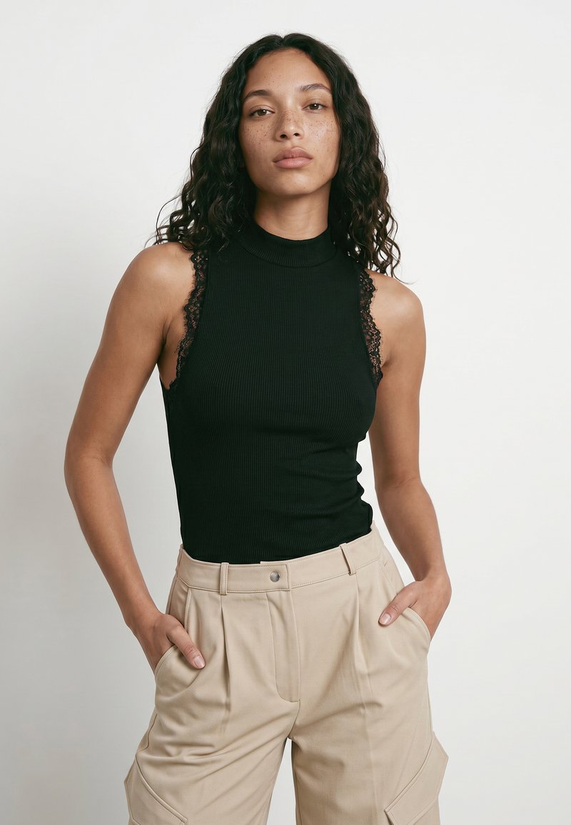 Femme aux cheveux bouclés foncés portant un top noir sans manches à col montant avec bordure en dentelle et un pantalon beige avec des poches, debout les mains dans les poches.