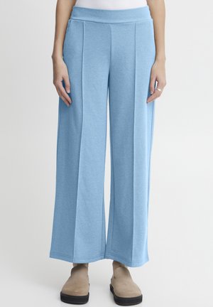 Woman wearing light blue wide-leg pants with front seams, beige slip-on shoes, and a white top, standing against a plain background.