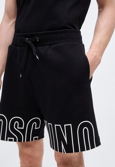 Άνδρας που φοράει μαύρα σορτς με κορδόνι και μεγάλα λευκά γράμματα "MOSCHINO" με περίγραμμα στα πόδια, στέκεται με το ένα χέρι στην τσέπη.