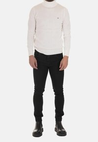 Man wearing a white textured turtleneck sweater, black slim-fit pants, and black leather boots standing against a plain background.