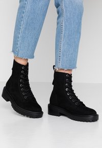 Botines de ante negro con cordones, suela de goma gruesa y punta redondeada. Presentan ojales de metal y una lengüeta en la parte posterior.