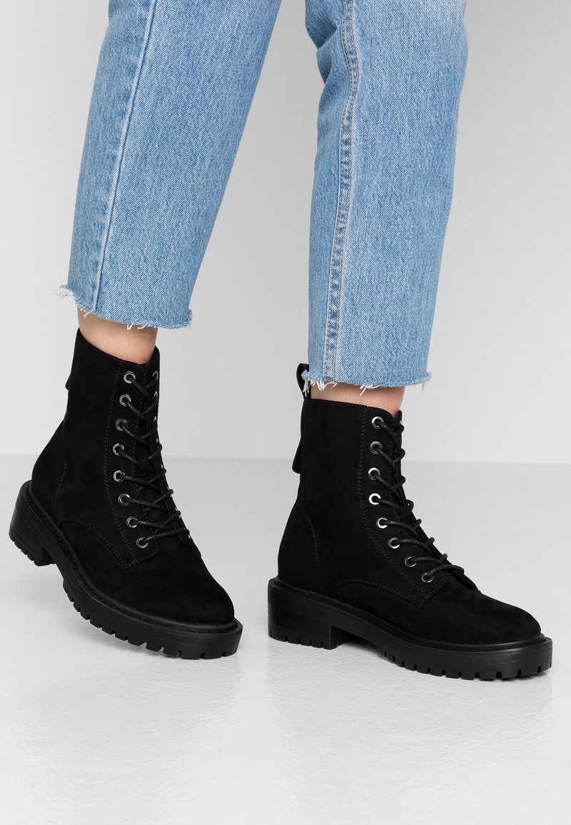 Botines de ante negro con cordones, suela de goma gruesa y punta redondeada. Presentan ojales de metal y una lengüeta en la parte posterior.