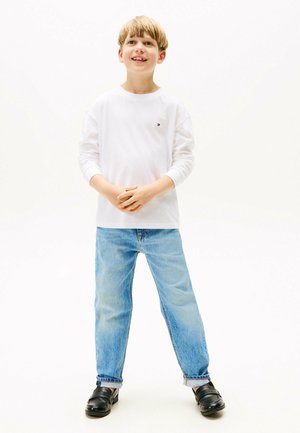 Niño pequeño de pie con las manos entrelazadas, vistiendo una camisa blanca de manga larga, vaqueros azul claro y zapatos negros sobre un fondo blanco liso.