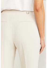 Beiga byxor med en slät textur, med en krok- och öglestängning och två bakfickor, visade från en bakifrån-perspektiv.