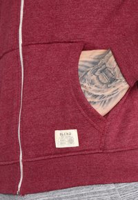 Rød zip-up hettegenser med v-hals, med en etikett som leser "BLEND." Viser en frontlomme og synlig tatovering på en hånd.