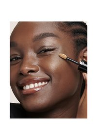 Applicateur de correcteur avec une pointe en éponge, tenu contre la peau. Modèle avec une peau lisse et foncée montrant un éclat naturel et un large sourire.