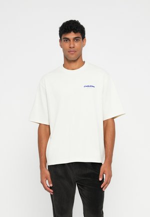 Witte katoenen t-shirt met korte mouwen, ronde halslijn, voorzien van blauwe tekst op de linkerborst. Draagt met donkere corduroy broek.