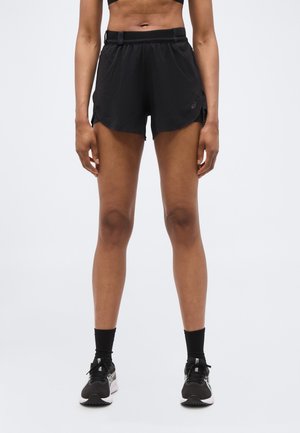 Αθλητής που φοράει μαύρο αθλητικό σορτς, μαύρες κάλτσες και μαύρα παπούτσια για τρέξιμο, στέκεται μπροστά σε απλό λευκό φόντο.