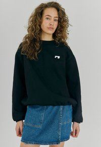 Sudadera negra de gran tamaño con un pequeño logo blanco en el pecho, combinada con una mini falda de mezclilla azul clara que tiene bolsillos frontales.