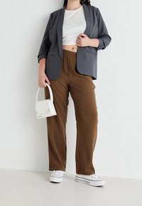 Šedý blejzr, bílý cropped top, hnědé pruhované kalhoty a bílé tenisky. Drží malou bílou kabelku. Jednoduchý design a uvolněný střih.