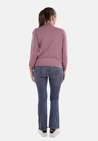 Donna con capelli castani raccolti in una coda di cavallo, che indossa un maglione rosa, jeans blu e sneakers bianche, in piedi rivolta verso l'indietro su uno sfondo bianco.
