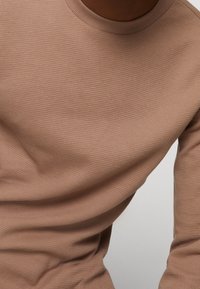 Waffle-Strick Langarmshirt in Hellbraun. Mit einem runden Halsausschnitt und strukturiertem Muster, mit einer taillierten Passform, die den Oberkörper betont.