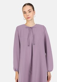 Pitkähihainen lilac-mekko, jossa on pyöreä pääntie, edessä nauhakoriste ja joustavat hihansuut. Mekko on valmistettu kevyestä, teksturoidusta kankaasta.