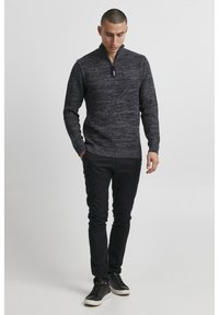 Mørk grå strikkegenser med høy krage og halv glidelås. Brukt sammen med svarte slim-fit bukser og svarte joggesko med hvite såler.