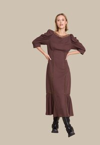 Brun langermet kjole med teksturert stoff, blondehals, og lagdelt kant, med vertikale dekorative sømmer. Brukt med svarte støvler.