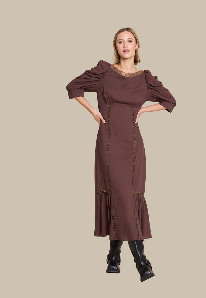 Brun langermet kjole med teksturert stoff, blondehals, og lagdelt kant, med vertikale dekorative sømmer. Brukt med svarte støvler.